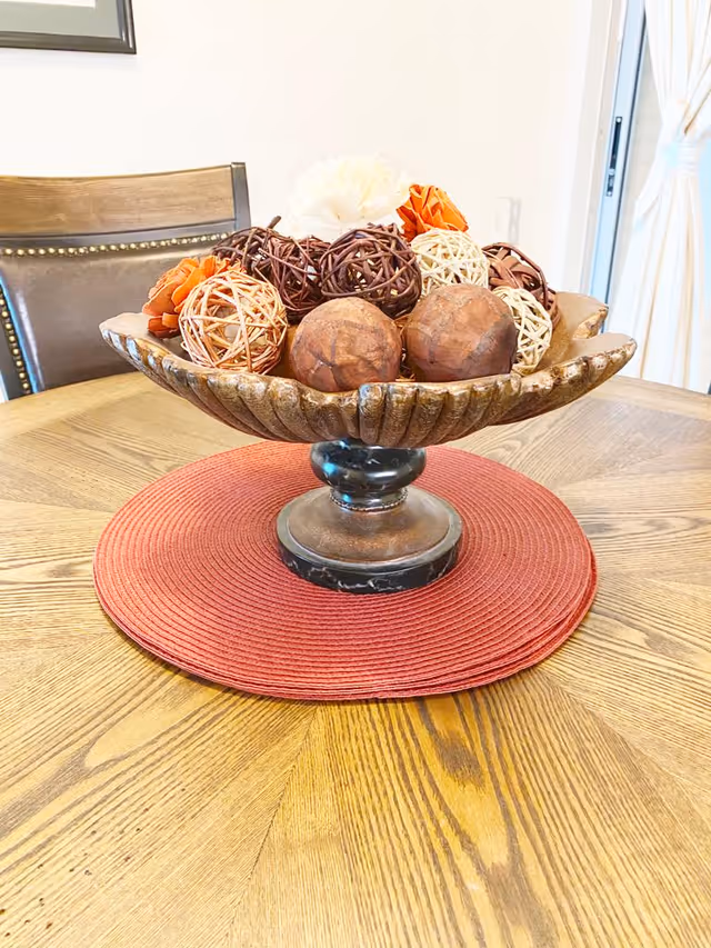 A decorative bowl filled with various textured and colored decorative balls, placed on a red circular placemat on a wooden table. In the background, there is a wooden chair with a leather backrest and a window with white curtains.