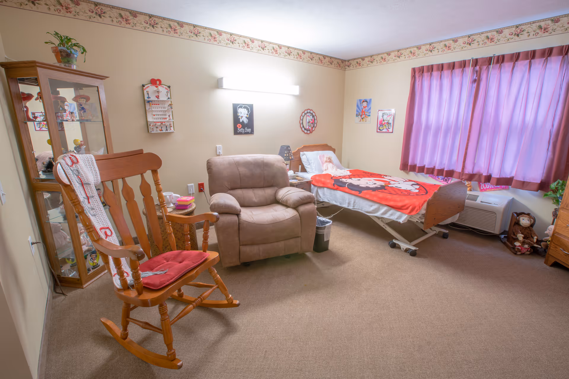 A cozy nursing home bedroom with a single bed covered by a red blanket featuring a cartoon character. Next to the bed is a beige recliner chair and a wooden rocking chair with a red cushion and a white crocheted blanket draped over it. The room has beige walls with a floral border near the ceiling, a glass display cabinet with collectibles, purple curtains covering the window, and various cartoon-themed decorations on the walls.