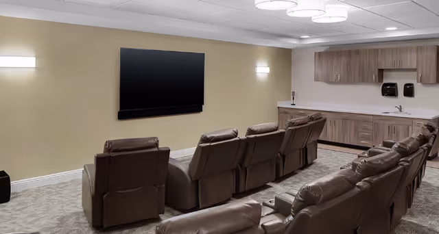 A cozy media room with two rows of brown leather recliners facing a large flat-screen TV mounted on a beige wall. The room has soft lighting from wall sconces and ceiling fixtures, and a kitchenette area with wooden cabinets, a sink, and soap dispensers is visible in the background.