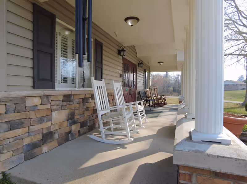 A covered outdoor porch area with white and brown rocking chairs lined up along a stone and siding wall. There are large white columns supporting the roof, and a red double door is visible in the background. The porch overlooks a grassy area with trees and a building in the distance.