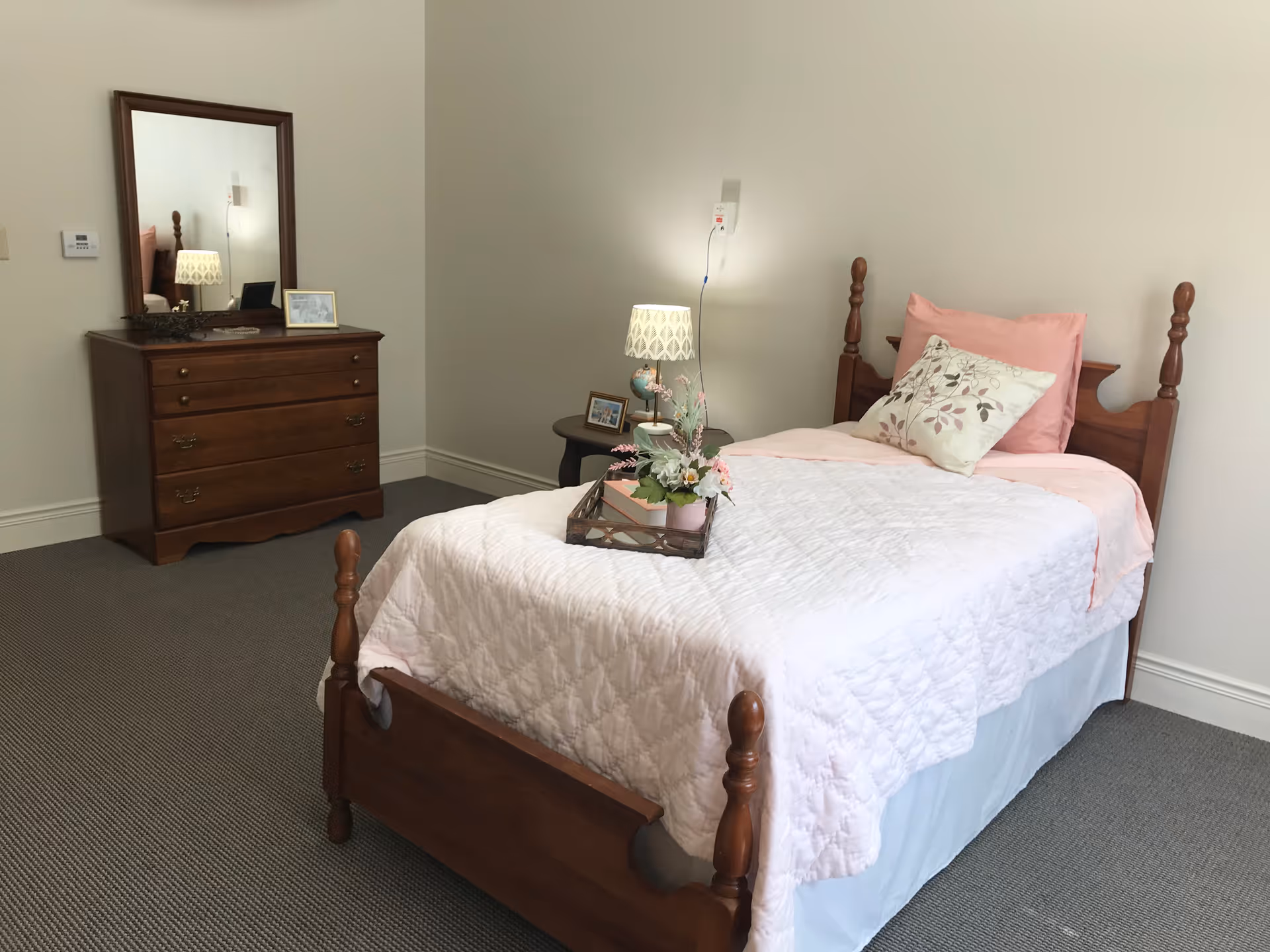 A neatly made single bed with a wooden frame, white quilt, pink pillow, and floral cushion in a simple bedroom. A wooden dresser with a mirror and a small table with a lamp and framed photos are visible in the background. The room has beige walls and carpeted floor.