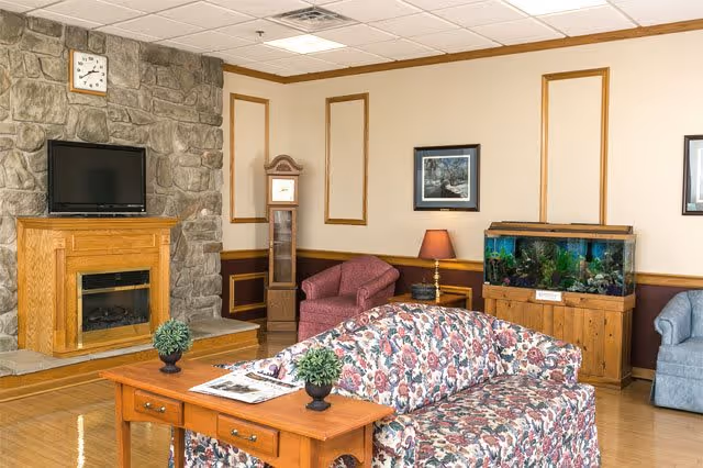 A cozy living room area with a floral patterned sofa facing a wooden coffee table with two small potted plants and a newspaper. Behind the sofa, there is a stone fireplace with a TV mounted above it and a clock on the stone wall. To the right, there is a red armchair, a wooden grandfather clock, a side table with a lamp, and a large fish tank on a wooden stand. The walls are decorated with framed pictures and wooden trim, and the floor is polished wood.