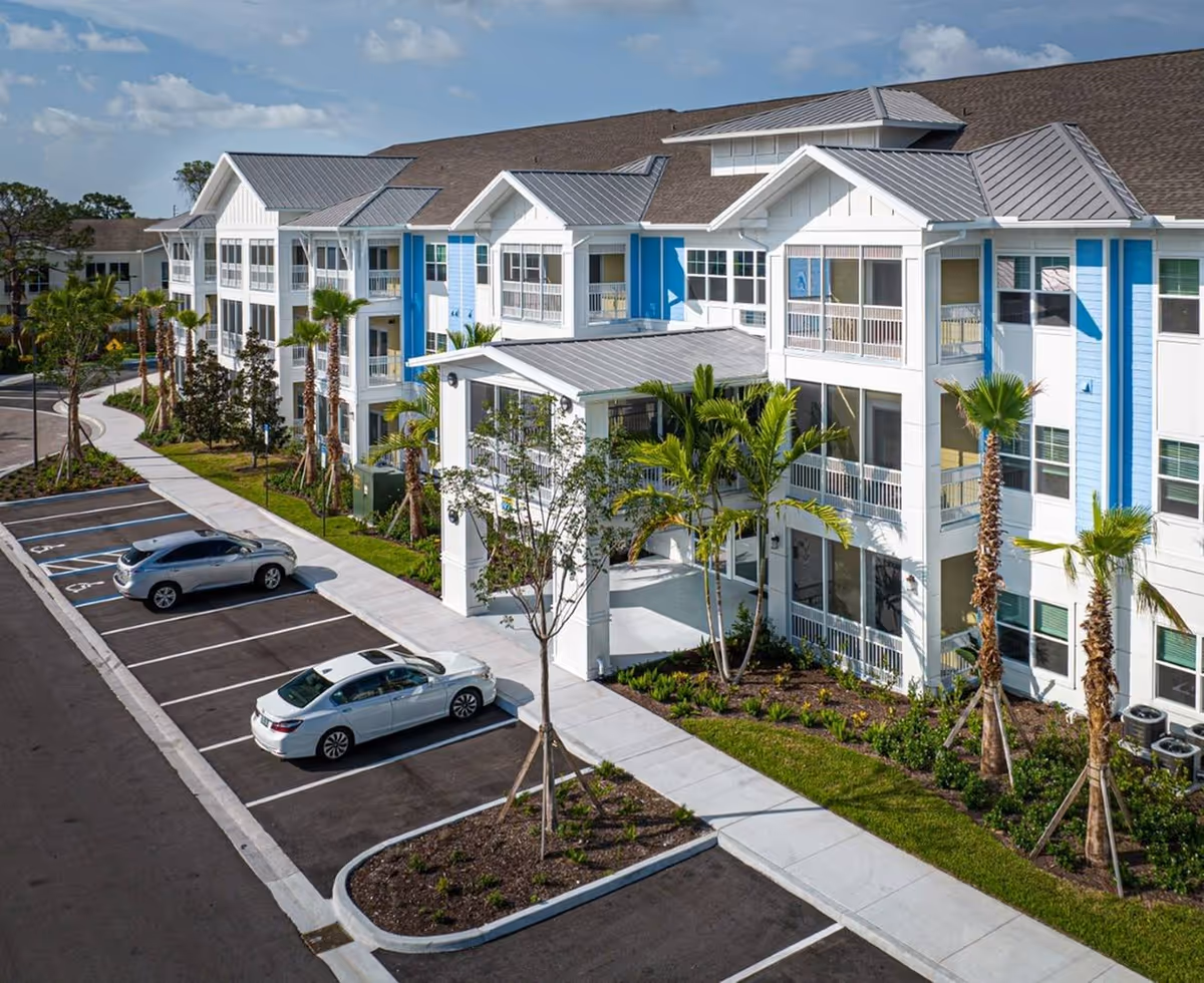 Exterior view of a three-story white and blue senior living building with balconies, palm trees, landscaping and a parking lot with cars.
