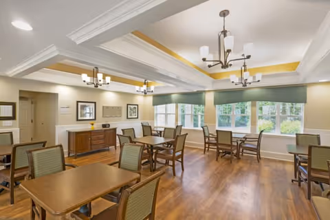A bright dining room with several wooden tables and chairs arranged neatly. The room features large windows with green valances allowing natural light to fill the space. The ceiling has decorative beams and modern chandeliers providing additional lighting. There is a wooden sideboard against one wall with framed artwork above it.