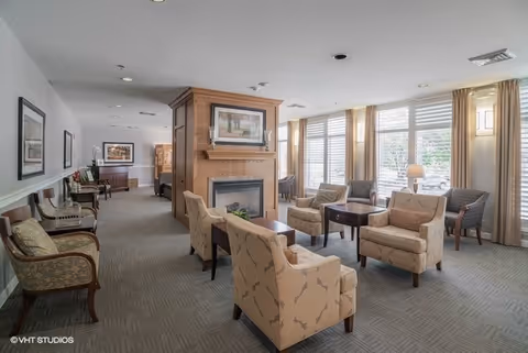 Well-lit senior living lounge with upholstered armchairs arranged around a central fireplace and large windows.