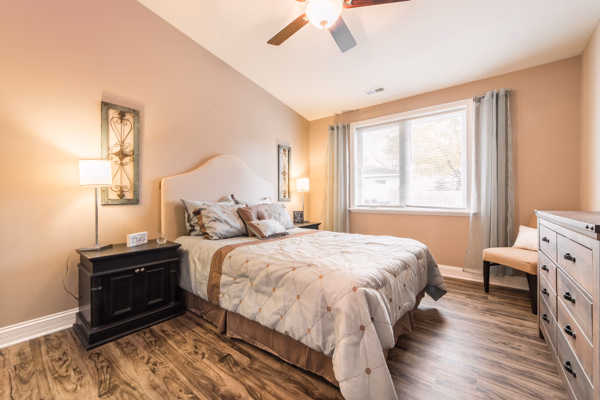 A cozy bedroom with a neatly made bed featuring a beige and brown quilt and multiple pillows. The room has light brown walls, a ceiling fan with a light, two bedside tables with lamps, decorative wall art, a window with blinds and light curtains, a wooden dresser, and a beige chair in the corner. The floor is wood with a natural finish.