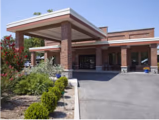 Exterior view of a healthcare and rehabilitation facility entrance with a covered drop-off area supported by brick columns, surrounded by landscaping with bushes and flowering plants under a clear blue sky.
