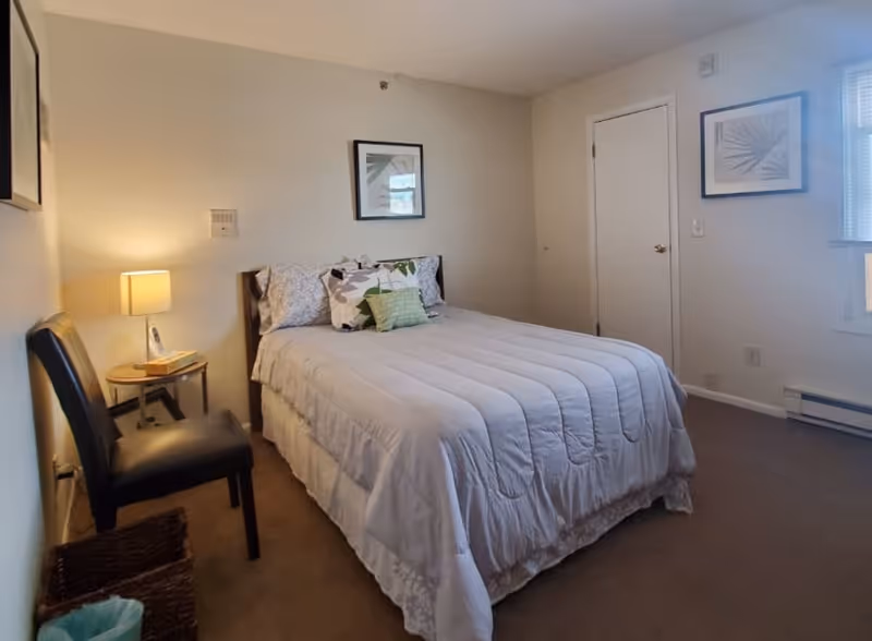 A bedroom with a neatly made bed covered with a white comforter and several decorative pillows. There is a black chair and a small round wooden side table with a lamp and a book on it. The walls are light-colored with two framed pictures hanging. A closed white door and a window with blinds are visible on the right side.