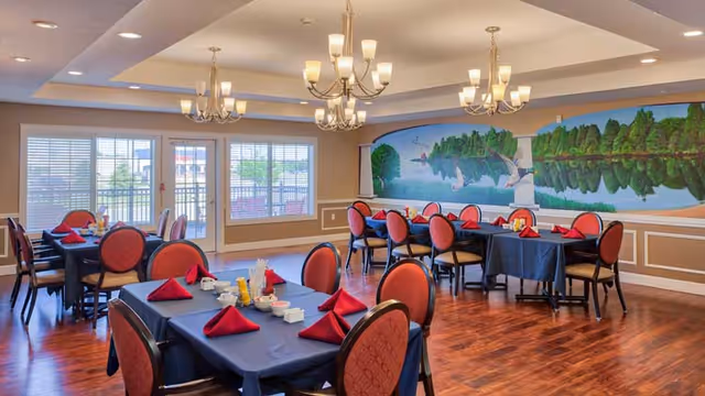Dining room with multiple tables covered in dark blue tablecloths and red napkins folded on each place setting. The room has wooden flooring, chandeliers hanging from the ceiling, large windows letting in natural light, and a mural of a lake scene with trees and birds on one wall.