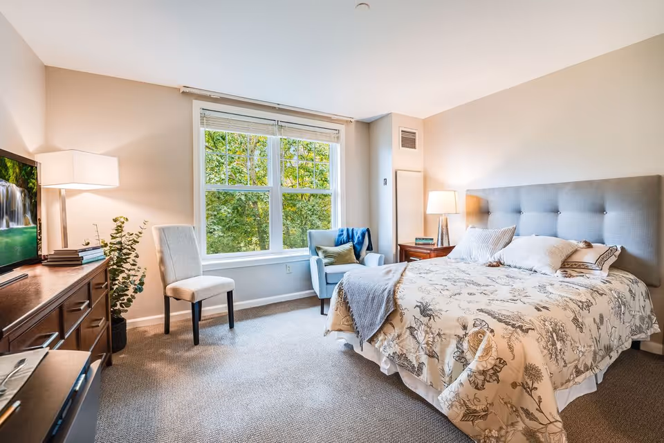 A bright and cozy bedroom with a large window showing green trees outside. The room features a bed with a floral patterned comforter and multiple pillows, a gray upholstered headboard, a wooden nightstand with a lamp and books, a light blue armchair with a green pillow and blue throw, a beige chair, a wooden dresser with a flat-screen TV displaying a waterfall, and a floor lamp next to the dresser.