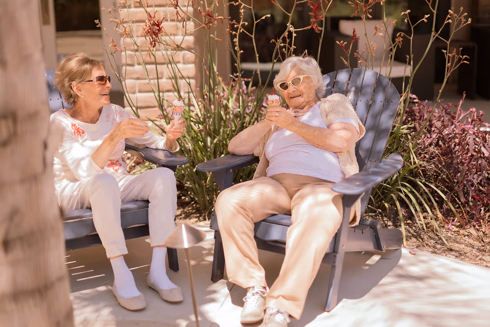 Two elderly women sitting outdoors in Adirondack chairs, enjoying ice cream cones and smiling at each other. They are surrounded by plants and sunlight, creating a warm and relaxed atmosphere.