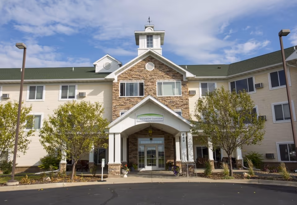 Front exterior view of Ecumen Countryside facility with a three-story building featuring beige siding and stone accents around the entrance. The entrance has a covered porch with white columns and a sign above the door that reads 'Ecumen Countryside'. There are trees and landscaping in front of the building and a paved driveway leading up to the entrance under a partly cloudy sky.
