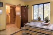 A small bedroom with a bed covered in a striped blanket, a wooden cabinet, and a window with blinds letting in natural light. The door to the room is open, revealing a bathroom with a shower and tiled walls.