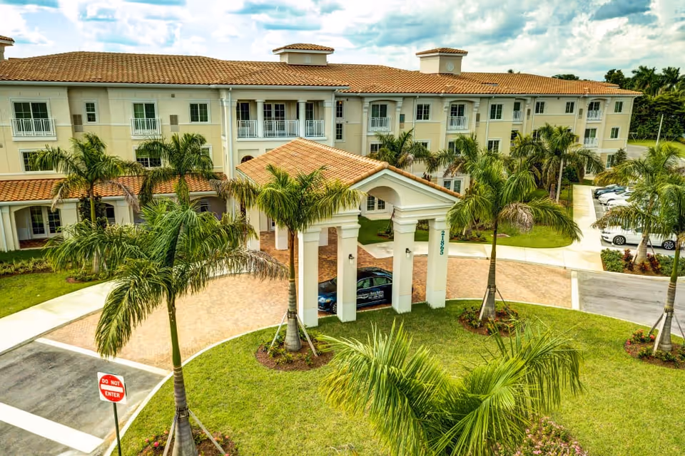 Exterior front of a three-story senior living building with a tiled porte-cochere, palm trees, and a circular driveway.