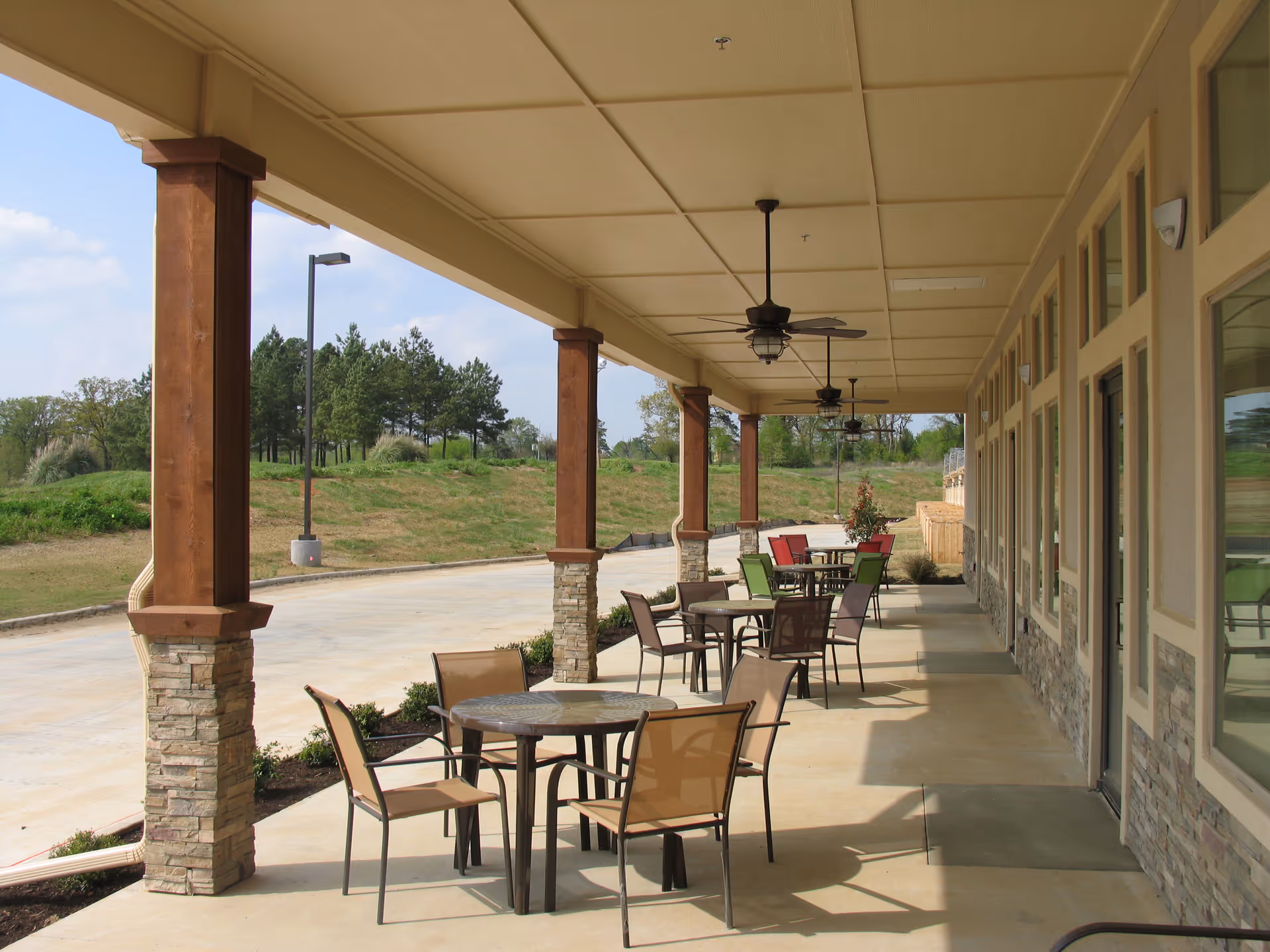 Covered outdoor patio area with multiple round tables and chairs arranged for seating. The patio has wooden pillars with stone bases, ceiling fans, and overlooks a grassy area with trees and a street lamp in the background.