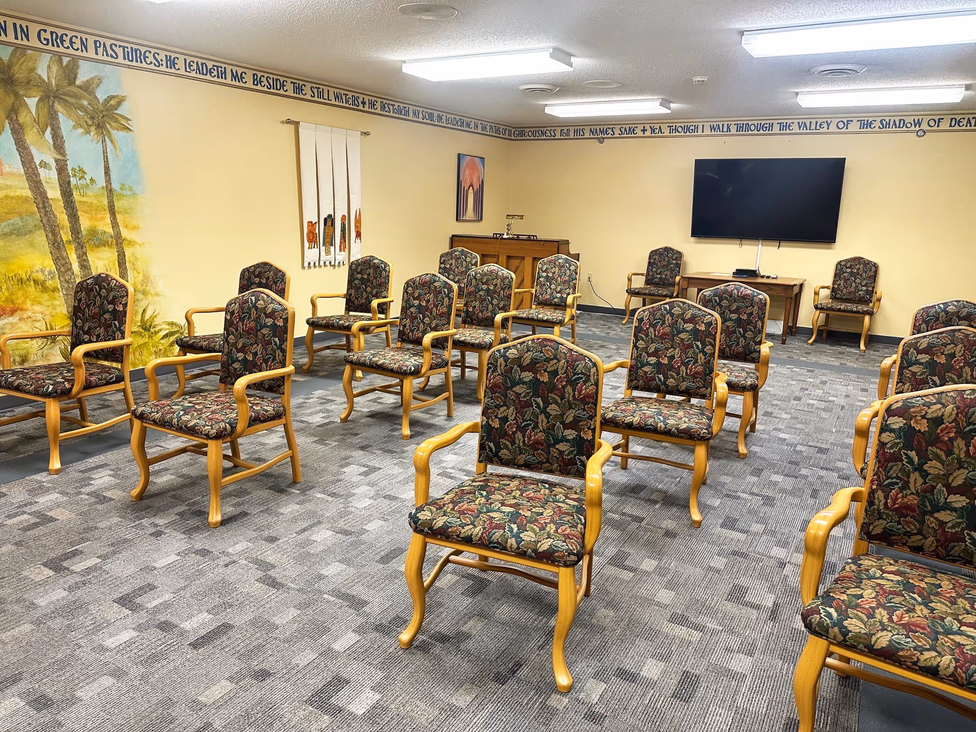 A room with multiple wooden chairs arranged in rows facing a wall-mounted flat screen TV. The chairs have floral patterned cushions. The walls are painted yellow and decorated with a mural of palm trees on one side and a religious-themed wall hanging and painting. There is a wooden cabinet beneath the TV and a carpeted floor with a gray patterned design. A scripture is written along the top edge of the walls near the ceiling.