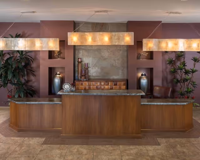 A reception desk area with a wooden counter and marble top, flanked by two large plants on either side. Behind the desk is a decorative wall with built-in shelves holding vases and candles. Three rectangular pendant lights hang from the ceiling, illuminating the space.