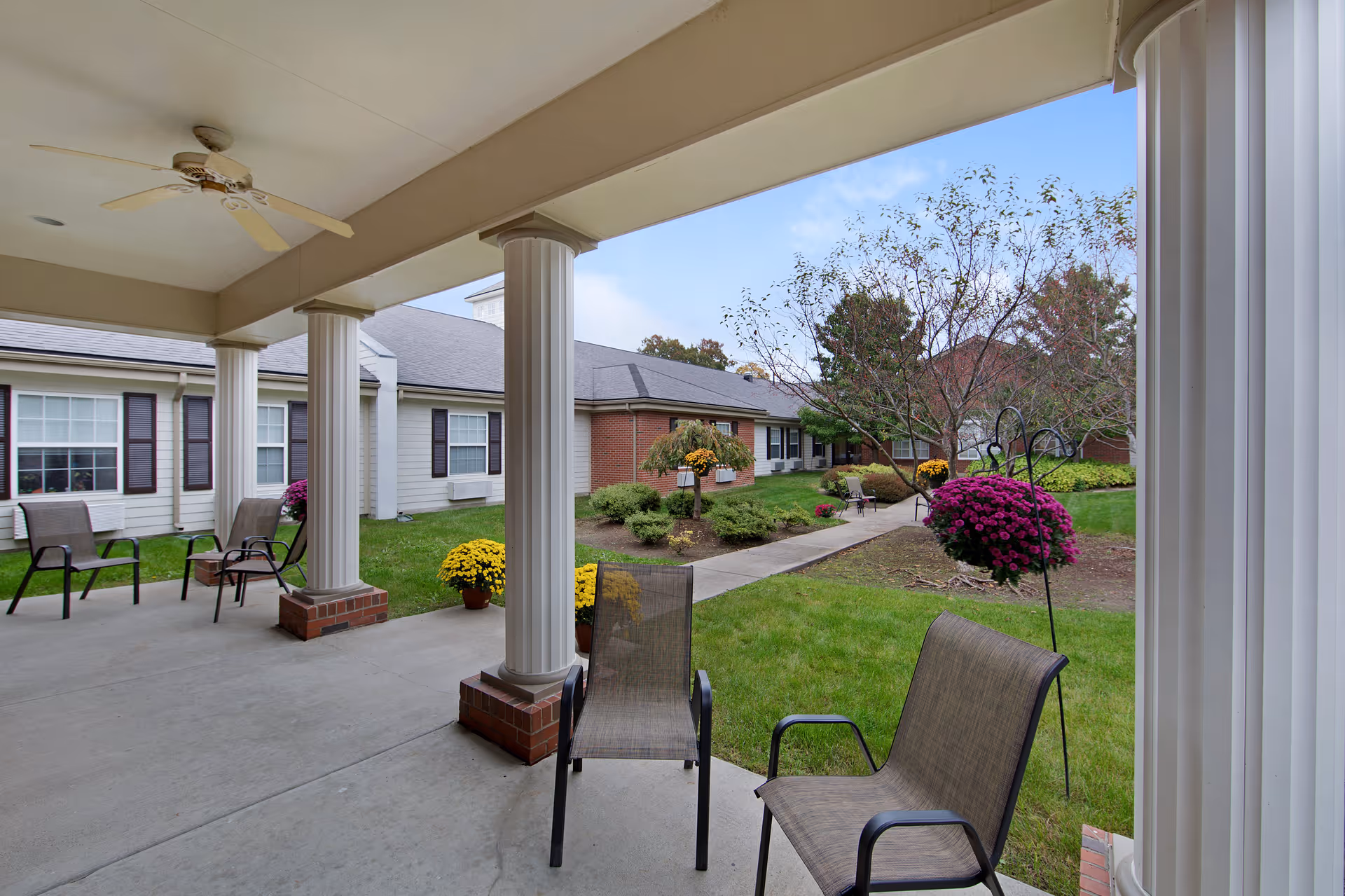 Covered outdoor patio area with several chairs and white columns overlooking a landscaped garden with green grass, shrubs, trees, and colorful flowers under a blue sky.