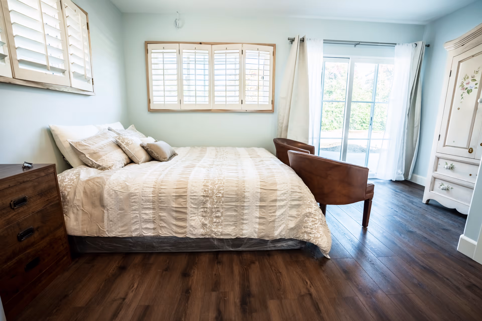 A bright bedroom with a large bed covered in a cream-colored comforter and multiple pillows. There are two brown chairs at the foot of the bed, a wooden dresser on the left, a white armoire with floral designs on the right, and a window with white shutters above the bed. Sliding glass doors with sheer white curtains lead to an outdoor area.