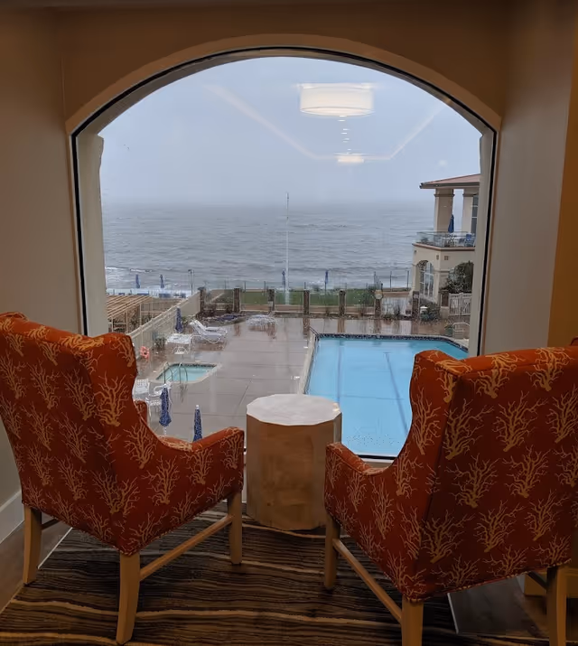 Two orange patterned chairs facing an arched window overlooking a pool and the ocean.