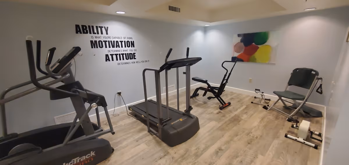 A small fitness room with exercise equipment including a treadmill, an elliptical machine, a rowing machine, and a pedal exerciser. The walls are light blue with a motivational quote decal that reads 'ABILITY is what you're capable of doing. MOTIVATION determines what you do. ATTITUDE determines how well you do it.' There is also a colorful abstract painting on one wall. The floor is wood-style laminate.