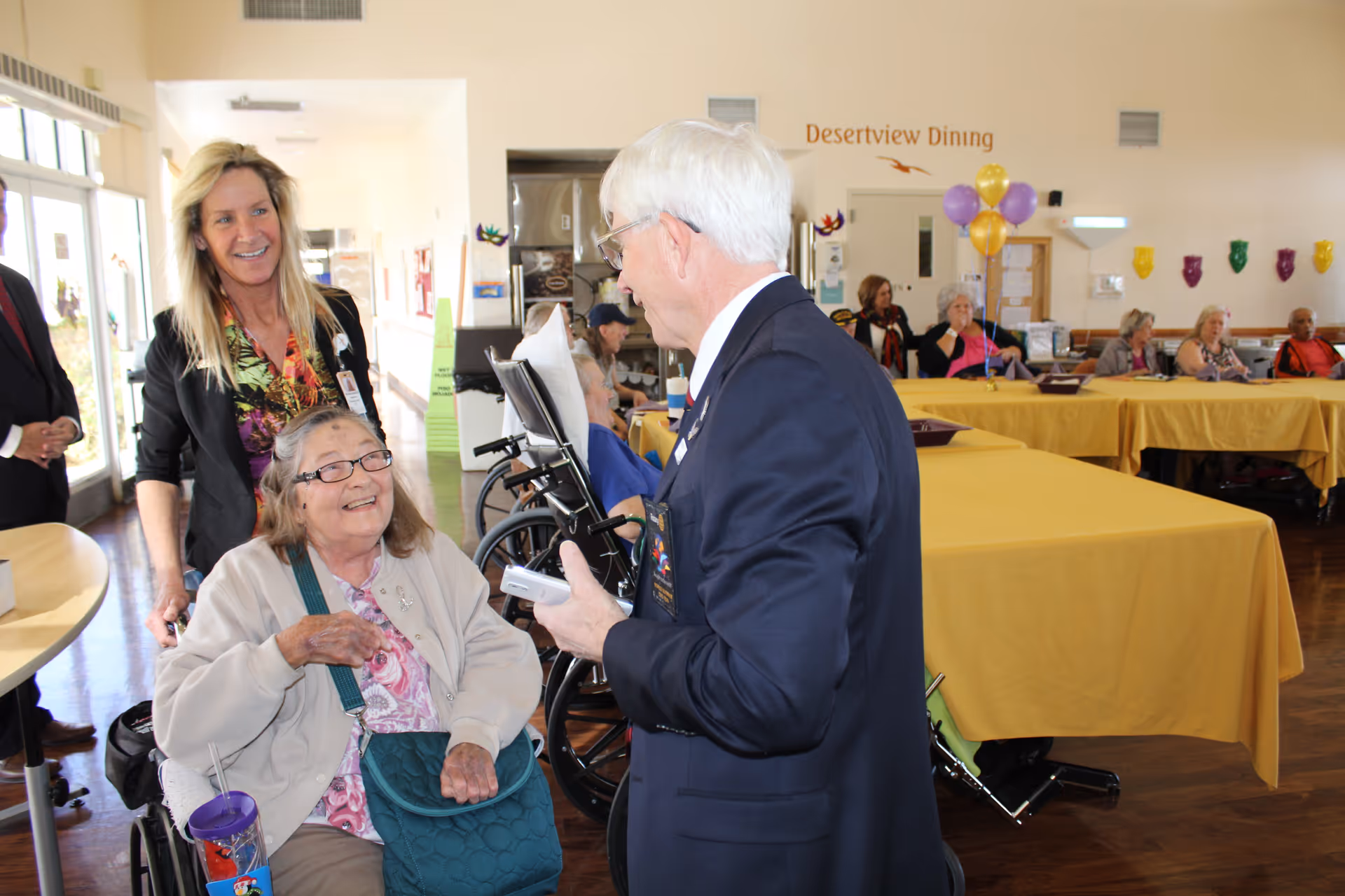 Residents and staff mingle in a bright dining room with tables covered in yellow tablecloths and a sign reading 'Desertview Dining'.