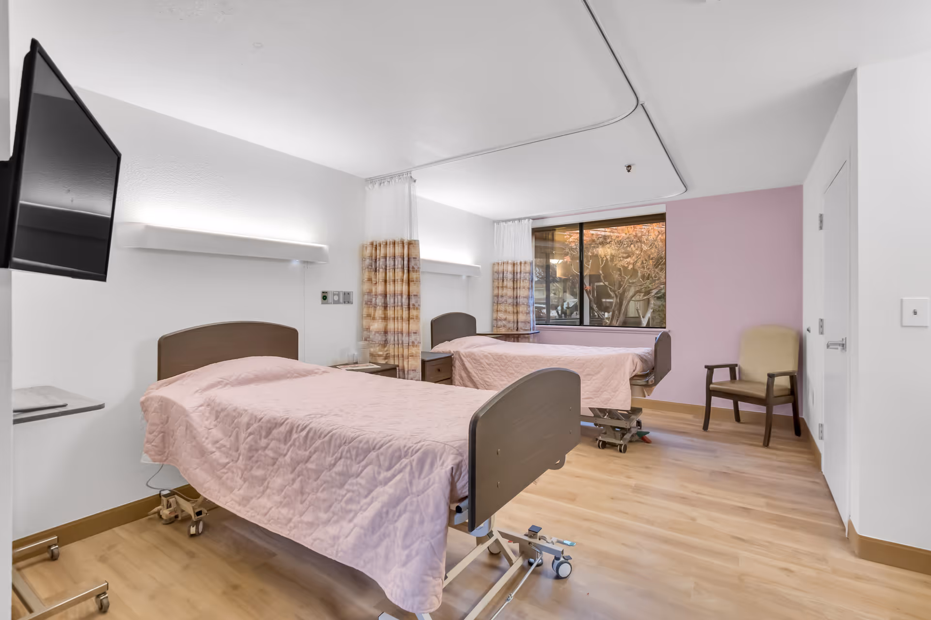 A clean and bright room with two hospital-style beds covered in light pink quilts. Each bed has a wooden headboard and is positioned near a window with patterned curtains. There is a beige chair in the corner, a wall-mounted TV, and light wood flooring. The walls are white with one accent wall painted light purple.