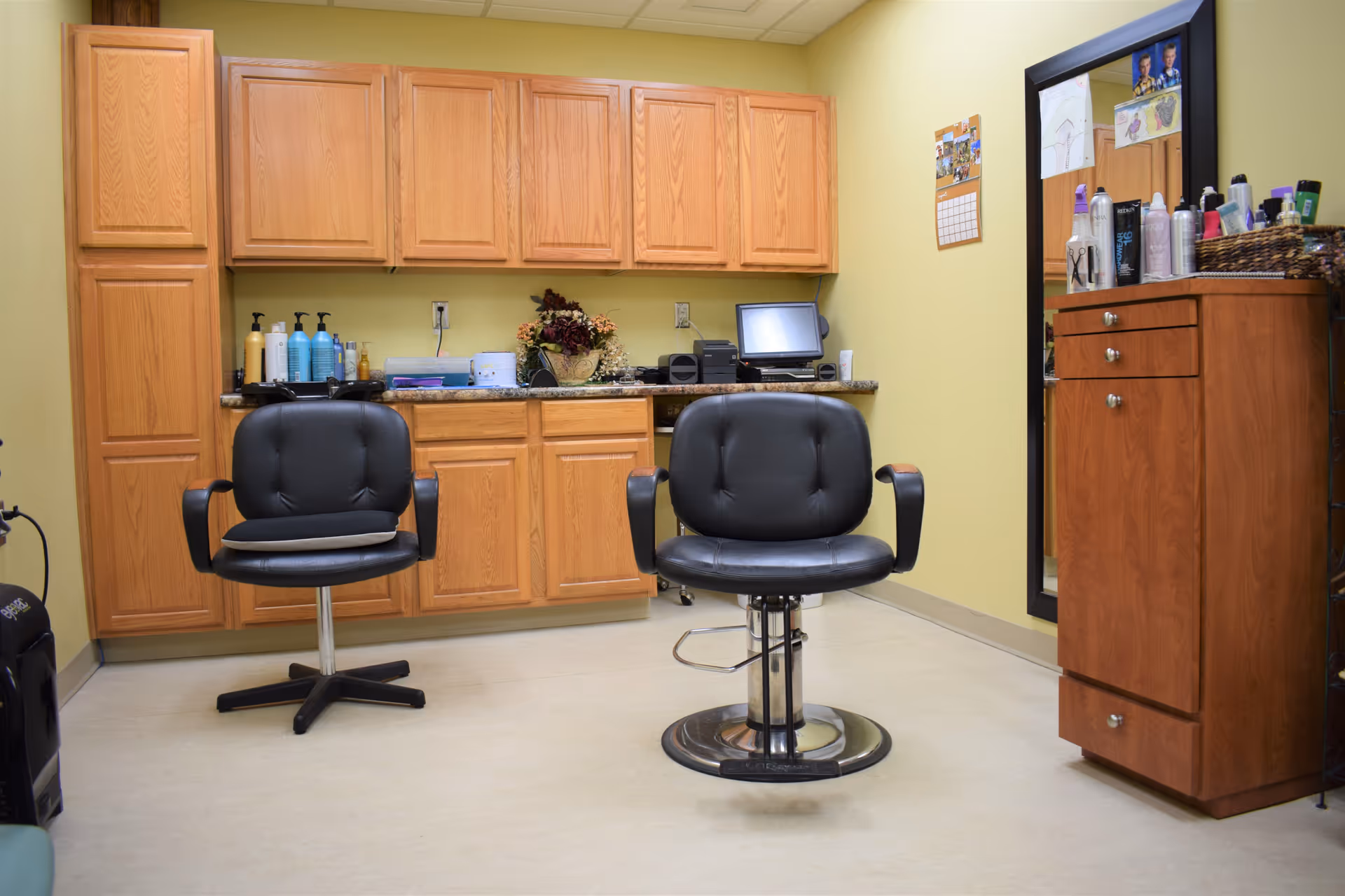 Interior view of a small salon or grooming room with two black salon chairs, wooden cabinets along the back wall, a countertop with various bottles and equipment, and a large mirror on the right wall with additional hair products on a wooden cabinet below it.