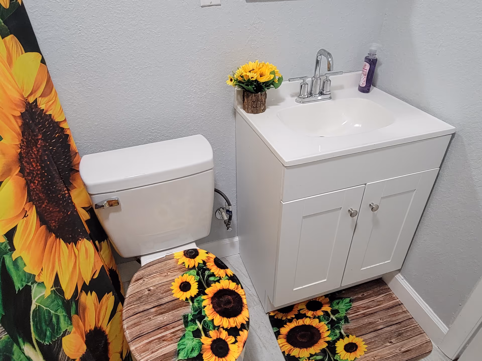 A small bathroom with a white toilet and a white vanity with a sink. The toilet seat cover, shower curtain, and floor mat all feature bright sunflower designs on a wooden plank background. A small vase with artificial sunflowers sits on the vanity next to a bottle of hand soap.