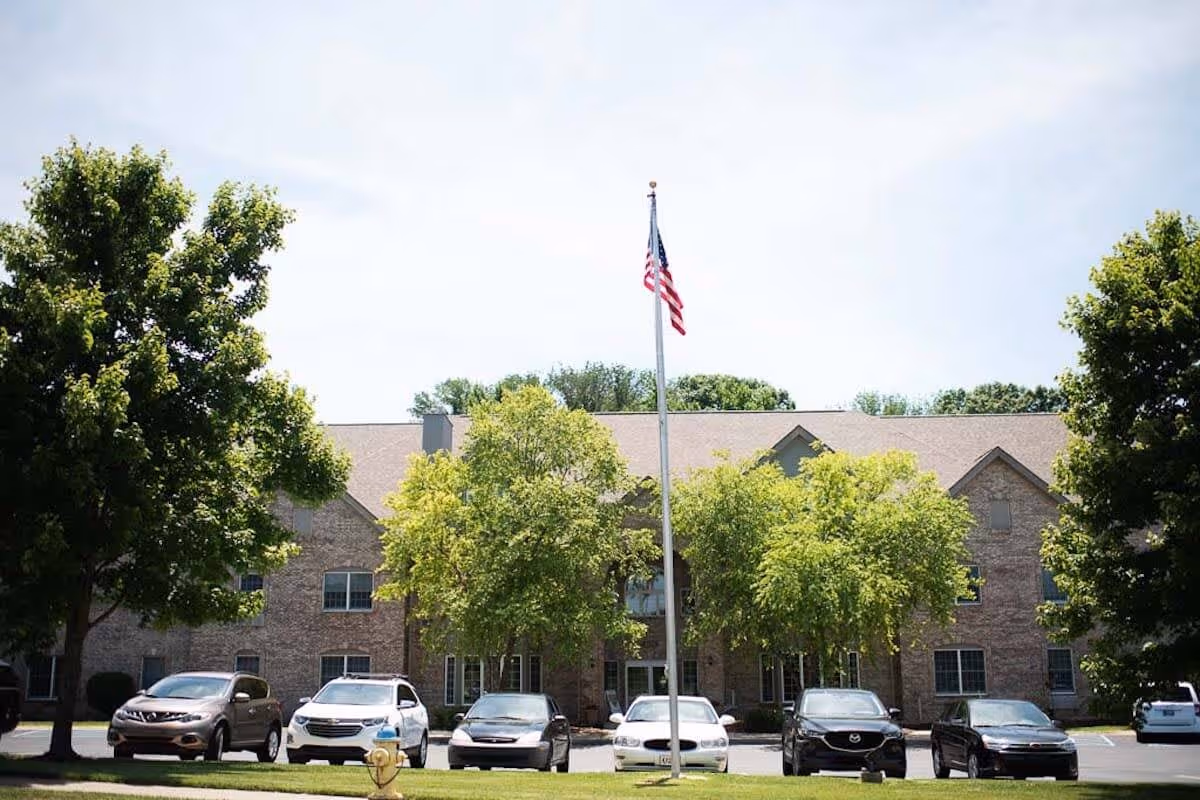 Exterior view of a two-story brick building with multiple windows, surrounded by green trees and a parking lot with several cars parked. An American flag on a flagpole is prominently displayed in front of the building under a clear sky.
