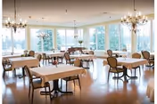 A spacious dining room with multiple tables covered with white tablecloths and surrounded by chairs. Large windows allow natural light to fill the room, and elegant chandeliers hang from the ceiling.
