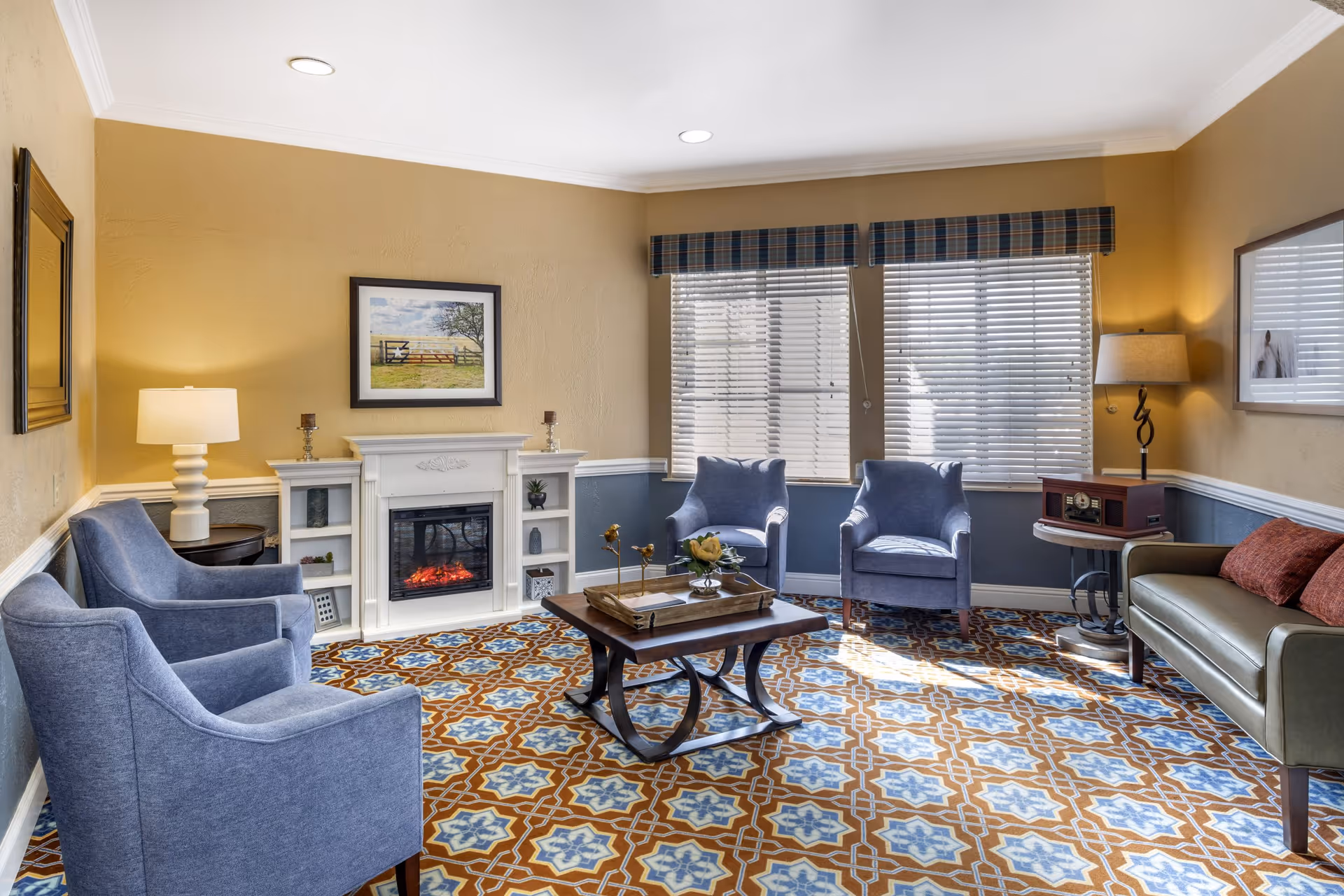 A cozy living room with four blue armchairs and a green sofa arranged around a wooden coffee table. The room features a patterned carpet with blue and brown designs, a white electric fireplace with shelves on either side, and two windows with blinds and plaid valances. There are two table lamps, one on a side table next to the sofa and another on a round table beside an armchair. The walls are painted beige with white trim, and framed artwork hangs above the fireplace and sofa.