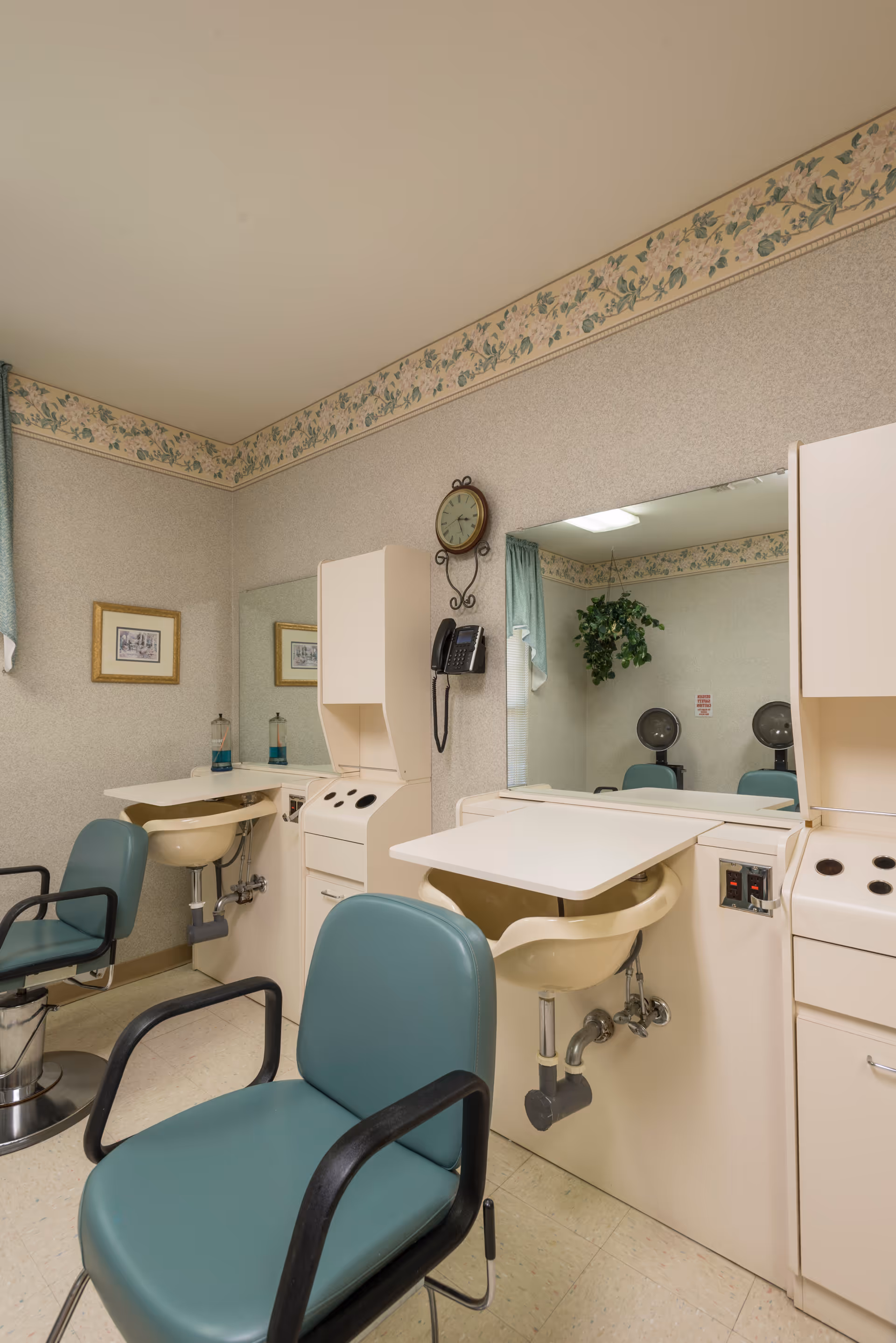 Interior view of a hair salon area in a skilled nursing facility featuring two green salon chairs in front of sinks and mirrors. The walls have floral wallpaper borders near the ceiling, and a clock and telephone are mounted on the wall. There is a framed picture and a potted plant visible in the reflection of the mirror.