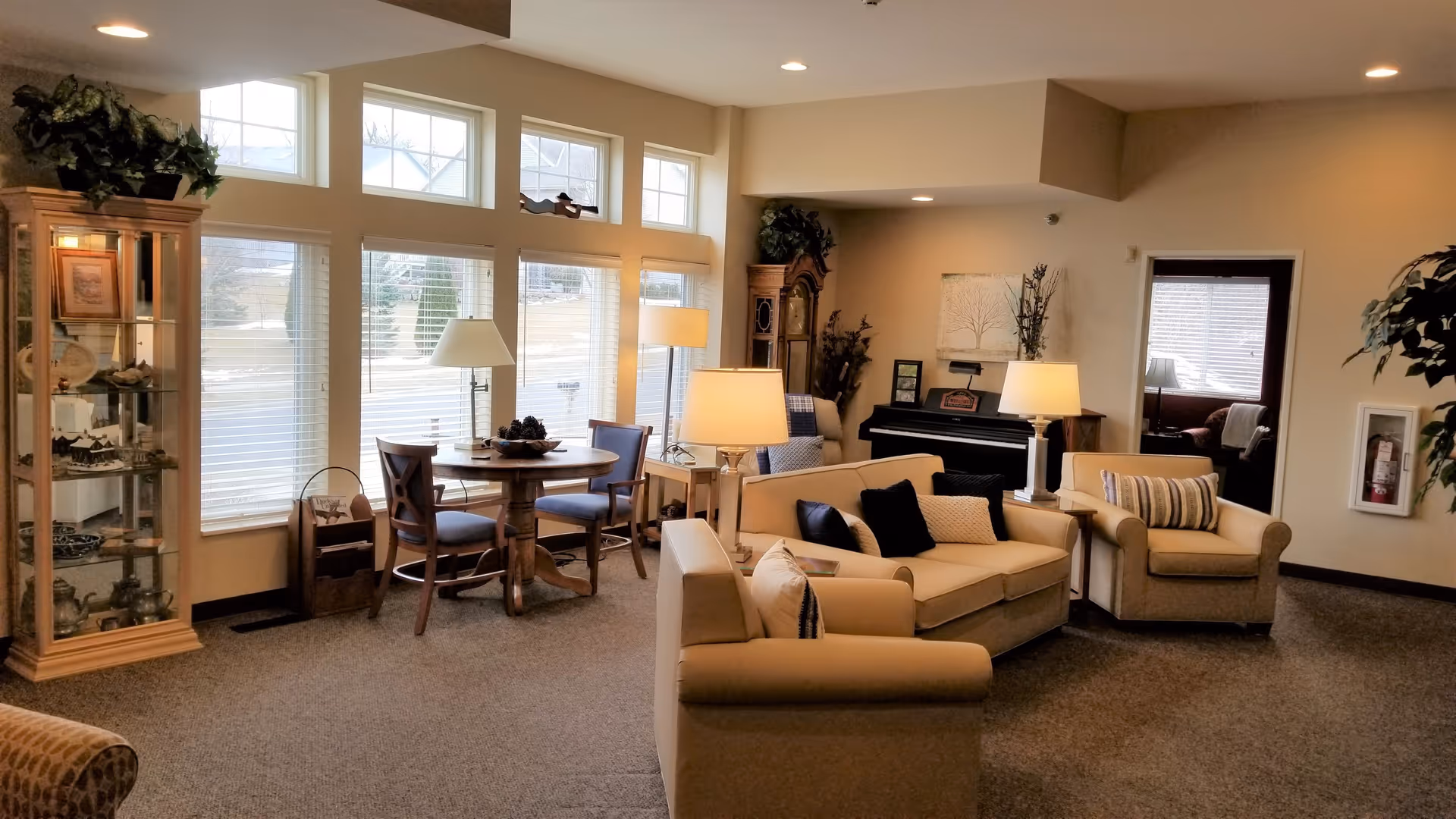 A cozy assisted living common area with beige sofas and armchairs arranged around a coffee table. There is a round wooden table with four chairs near large windows letting in natural light. The room also features a piano, floor lamps, a glass display cabinet with decorative items, and plants, creating a warm and inviting atmosphere.
