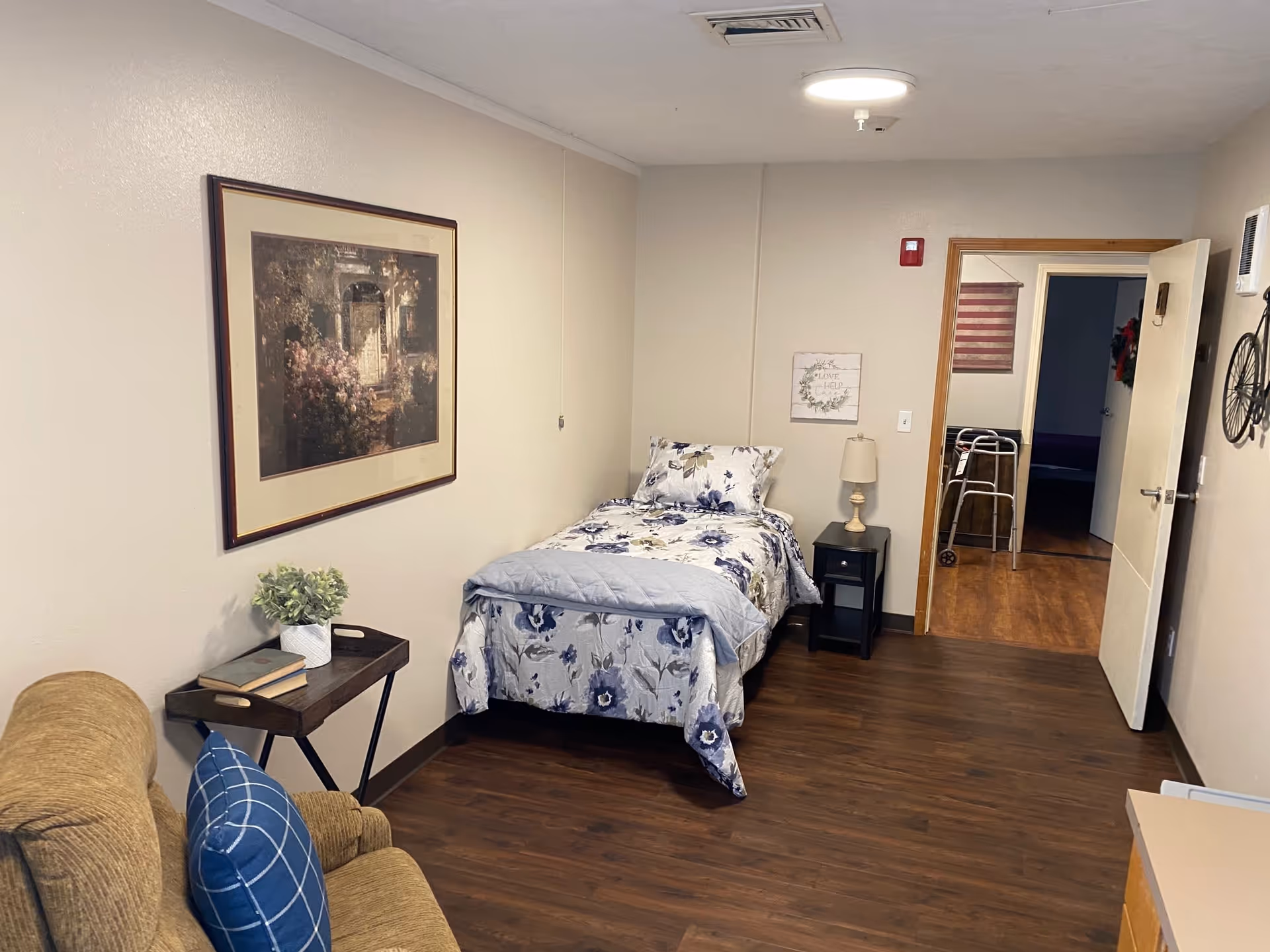 A small bedroom in a senior living facility featuring a single bed with floral bedding, a nightstand with a lamp, a framed picture on the wall, a brown armchair with a blue checkered pillow, a small table with books and a plant, and an open door showing a walker in the adjacent room.