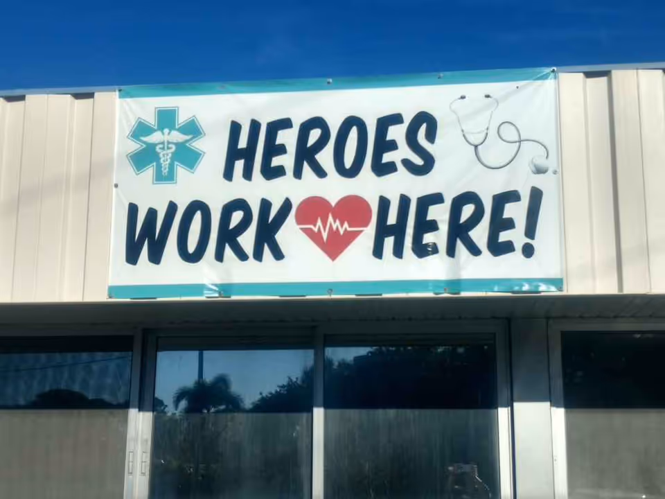 A banner on the exterior of a building with the text 'HEROES WORK HERE!' featuring a medical symbol, a heart with a heartbeat line, and a stethoscope graphic.