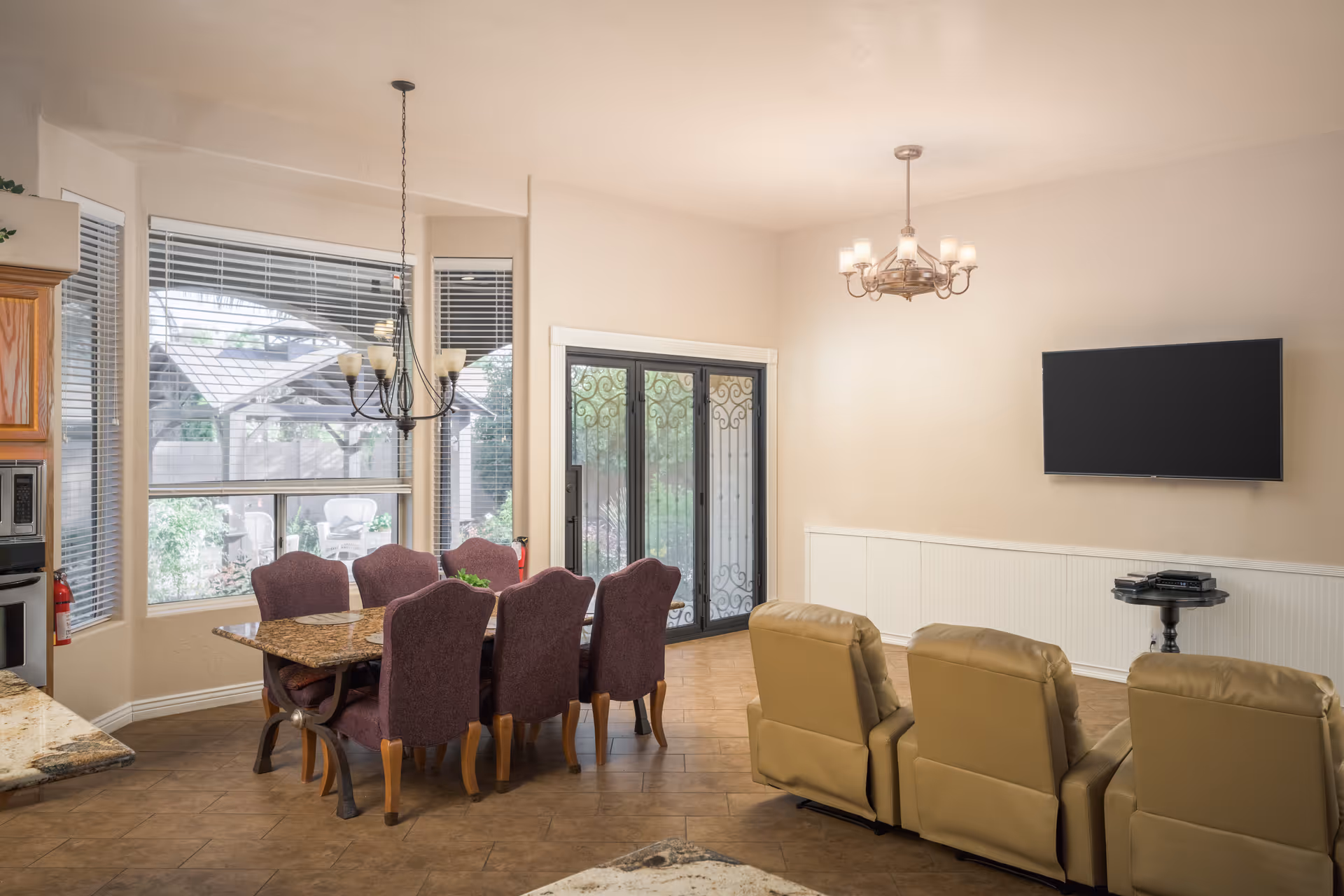 A bright and spacious interior room featuring a dining area with a rectangular table and six purple upholstered chairs, a living area with three beige recliners facing a wall-mounted flat-screen TV, large windows with blinds, and glass doors leading outside. The room has tiled flooring and two chandeliers hanging from the ceiling.