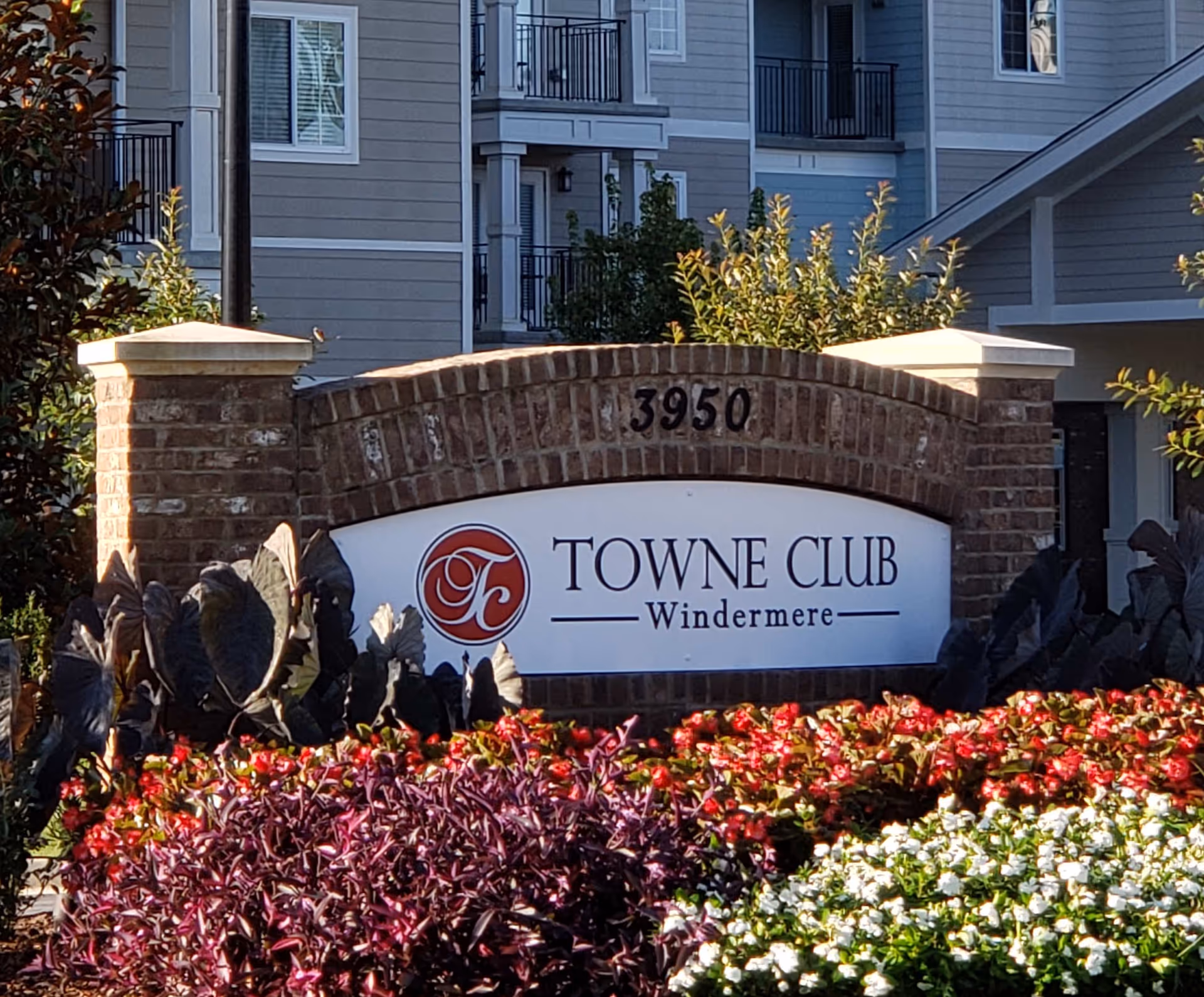 Brick sign with the address number 3950 and a white panel displaying the Towne Club Windermere logo and name, surrounded by colorful flowers and plants, with a residential building in the background.