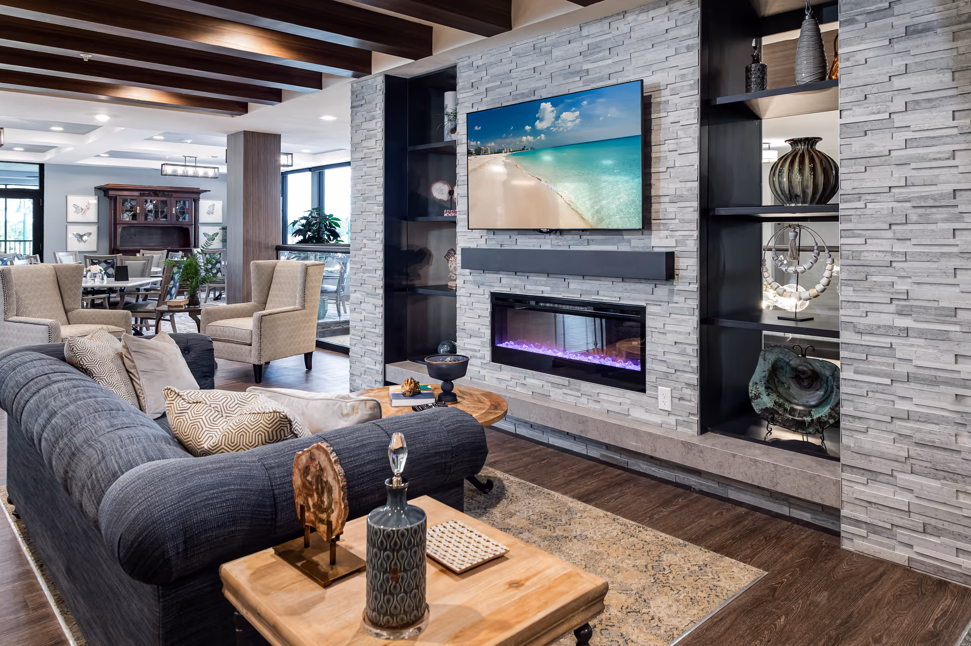 A modern living room area in a senior living facility featuring a dark gray sofa with patterned cushions, two beige armchairs, a wooden coffee table, and a wall-mounted flat-screen TV above a sleek electric fireplace. The room has a stone accent wall with built-in shelves displaying decorative items, wooden ceiling beams, and a dining area visible in the background with tables, chairs, and a wooden cabinet.
