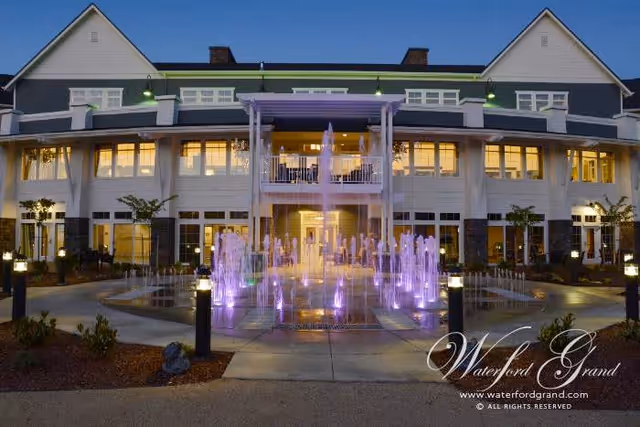 Exterior view of a large, well-lit senior living facility building at dusk with a central water fountain illuminated with purple lights in front. The building has multiple windows, a balcony, and a peaked roof.