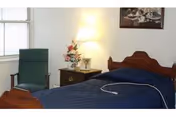 Single-bed bedroom with a blue quilt and wooden headboard, nightstand with a lamp and flowers, and a green chair by a window.