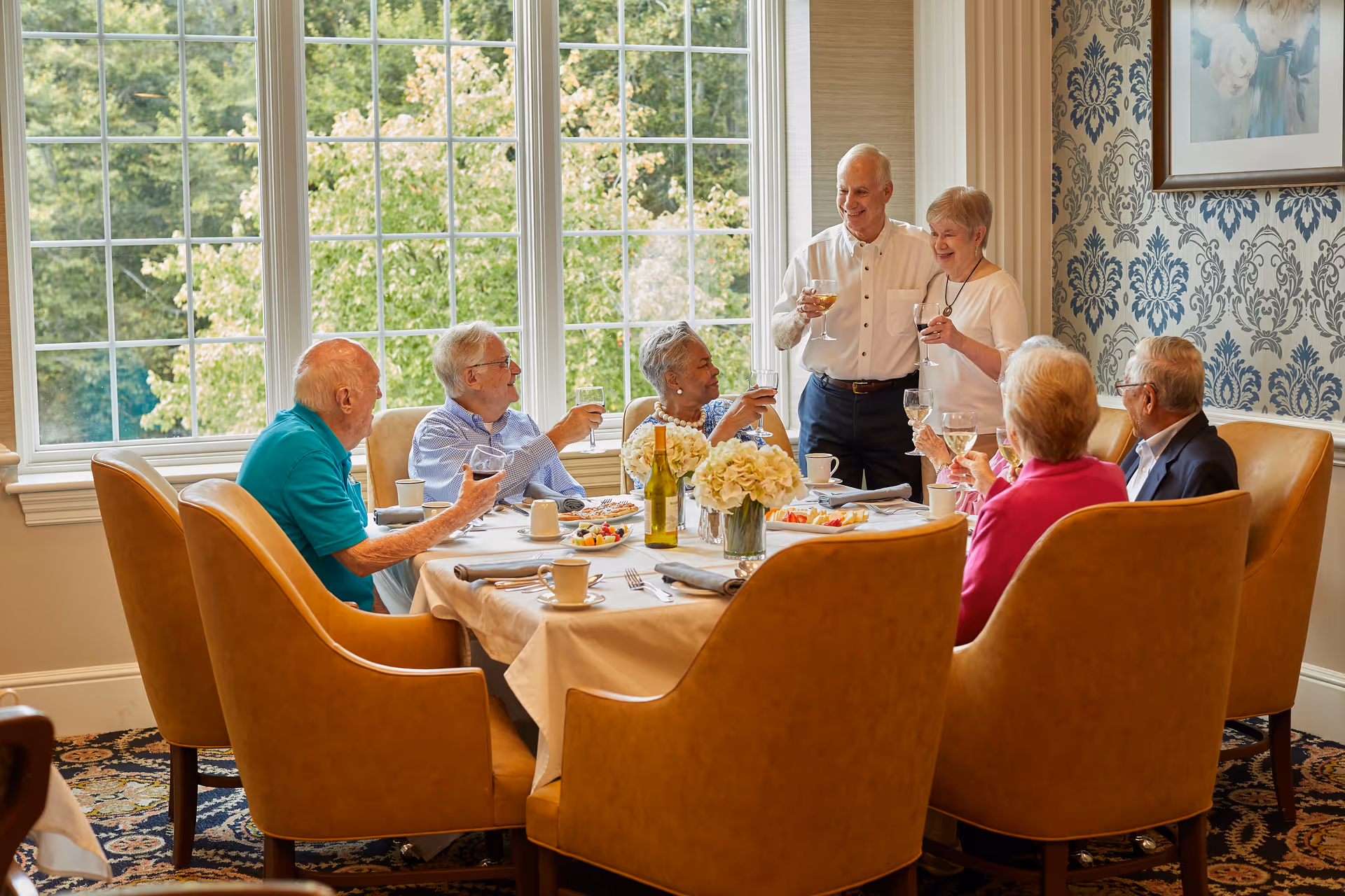 A group of seven elderly people sitting and standing around a dining table in a well-lit room with large windows and patterned wallpaper. They are raising their glasses in a toast, smiling and enjoying each other's company. The table is set with plates of food, cups, and a vase of flowers.