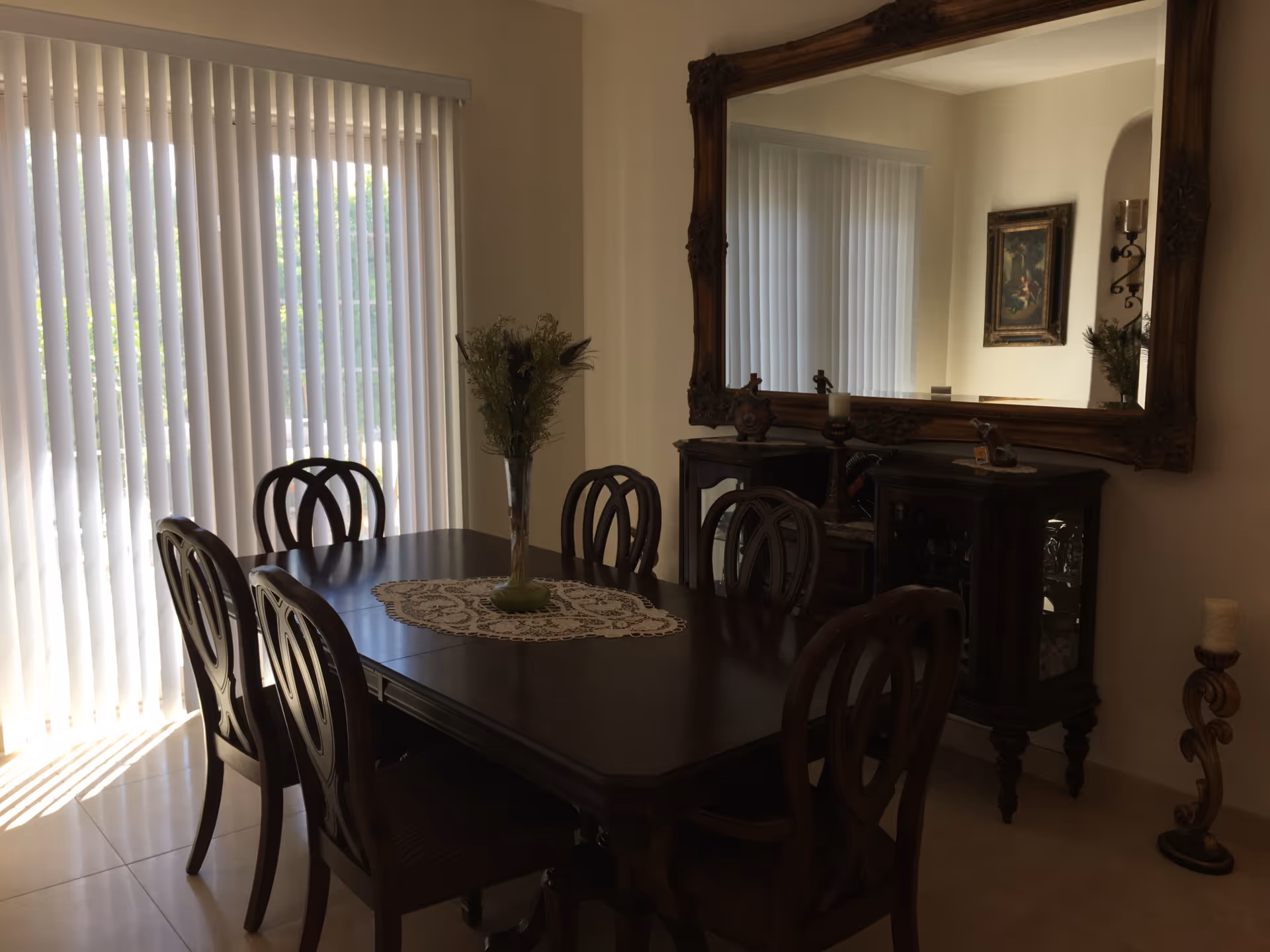 A dining room with a dark wooden table and six matching chairs. A lace table runner and a vase with flowers are placed on the table. Behind the table is a large ornate mirror hanging on the wall, reflecting part of the room. Vertical blinds cover a sliding glass door that lets in natural light.