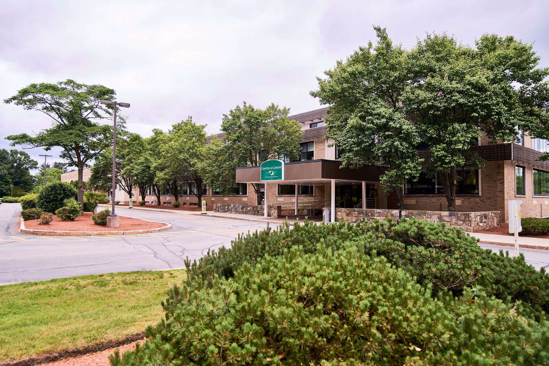 Exterior view of CareOne at Concord facility showing a two-story brick building surrounded by trees and landscaped bushes, with a driveway and parking area in front under a cloudy sky.