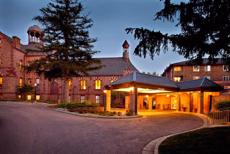 Twilight view of a brick senior living building with a covered, illuminated main entrance and tall trees.