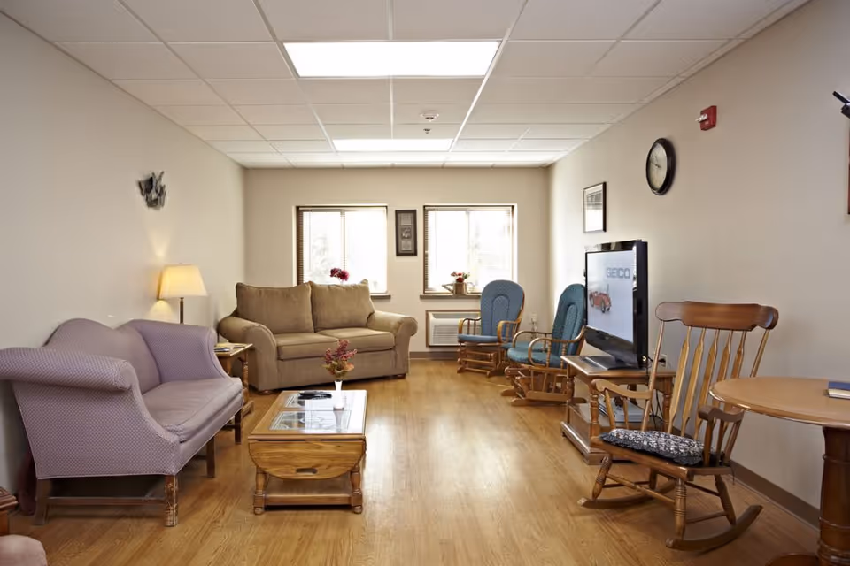A cozy living room in the River View facility featuring a beige sofa, a purple loveseat, two blue rocking chairs, a wooden rocking chair, a wooden coffee table with a small vase of flowers, a TV on a stand, and two windows letting in natural light.