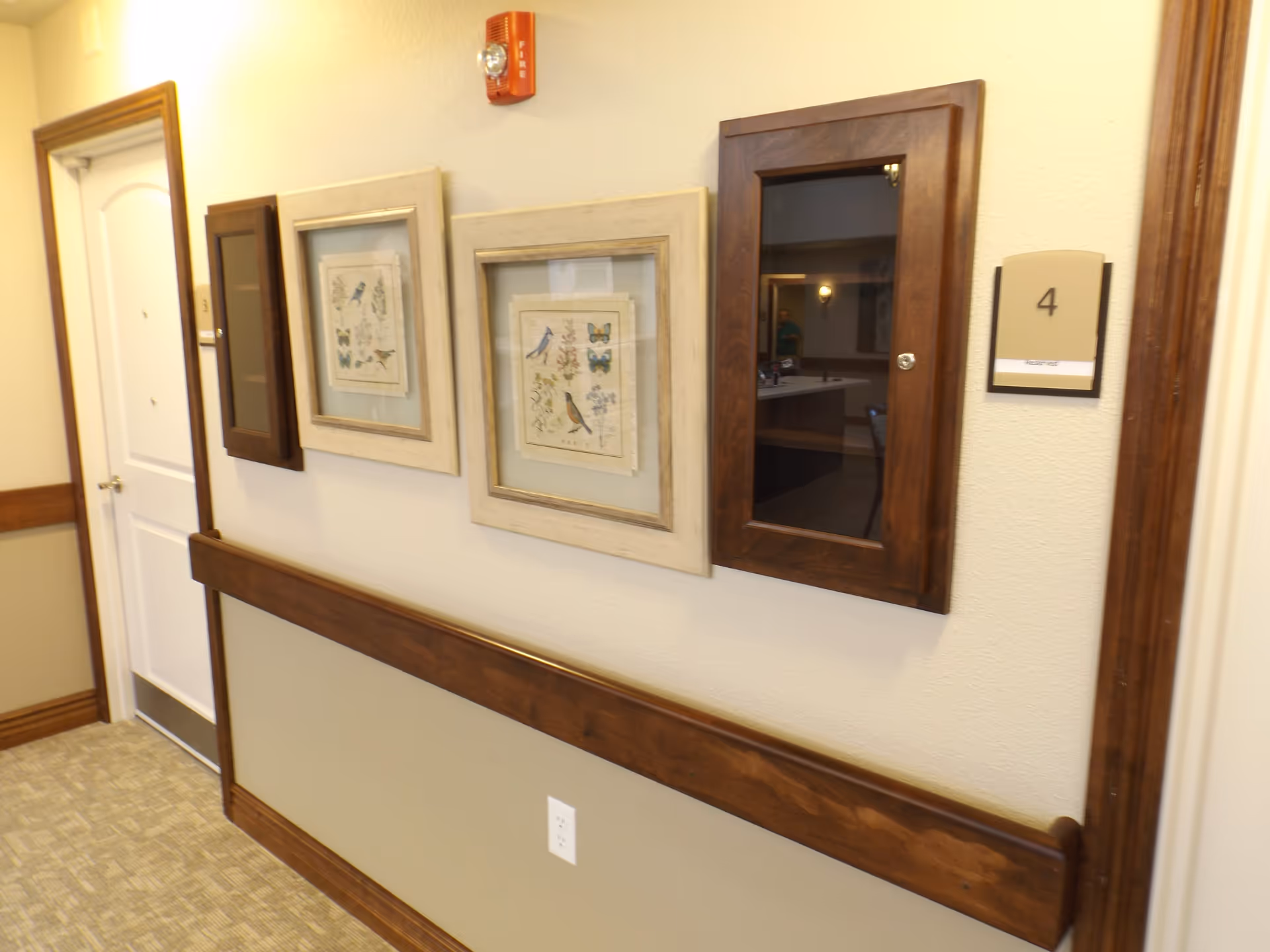 Interior hallway of a senior living facility with beige walls and carpeted floor. The wall features wooden trim and handrails, framed artwork of birds and butterflies, a fire alarm, and a wooden cabinet with a glass door. A door labeled with the number 4 is visible on the left side.