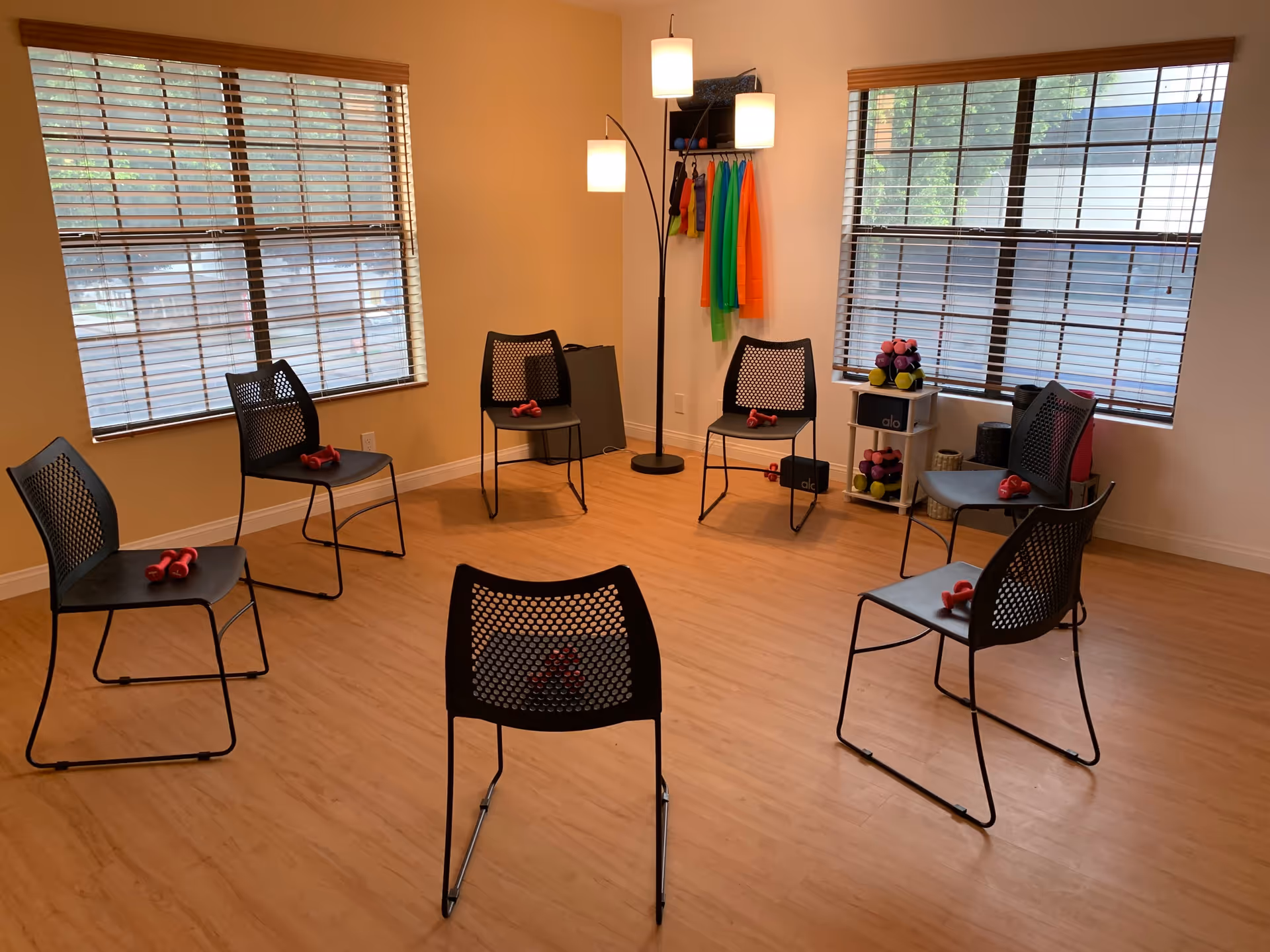A bright room with wooden floors and two large windows with blinds. Six black mesh chairs are arranged in a circle, each with a pair of small red dumbbells on the seat. In the corner, there is a tall floor lamp with three lights and a rack holding colorful exercise bands and small weights.