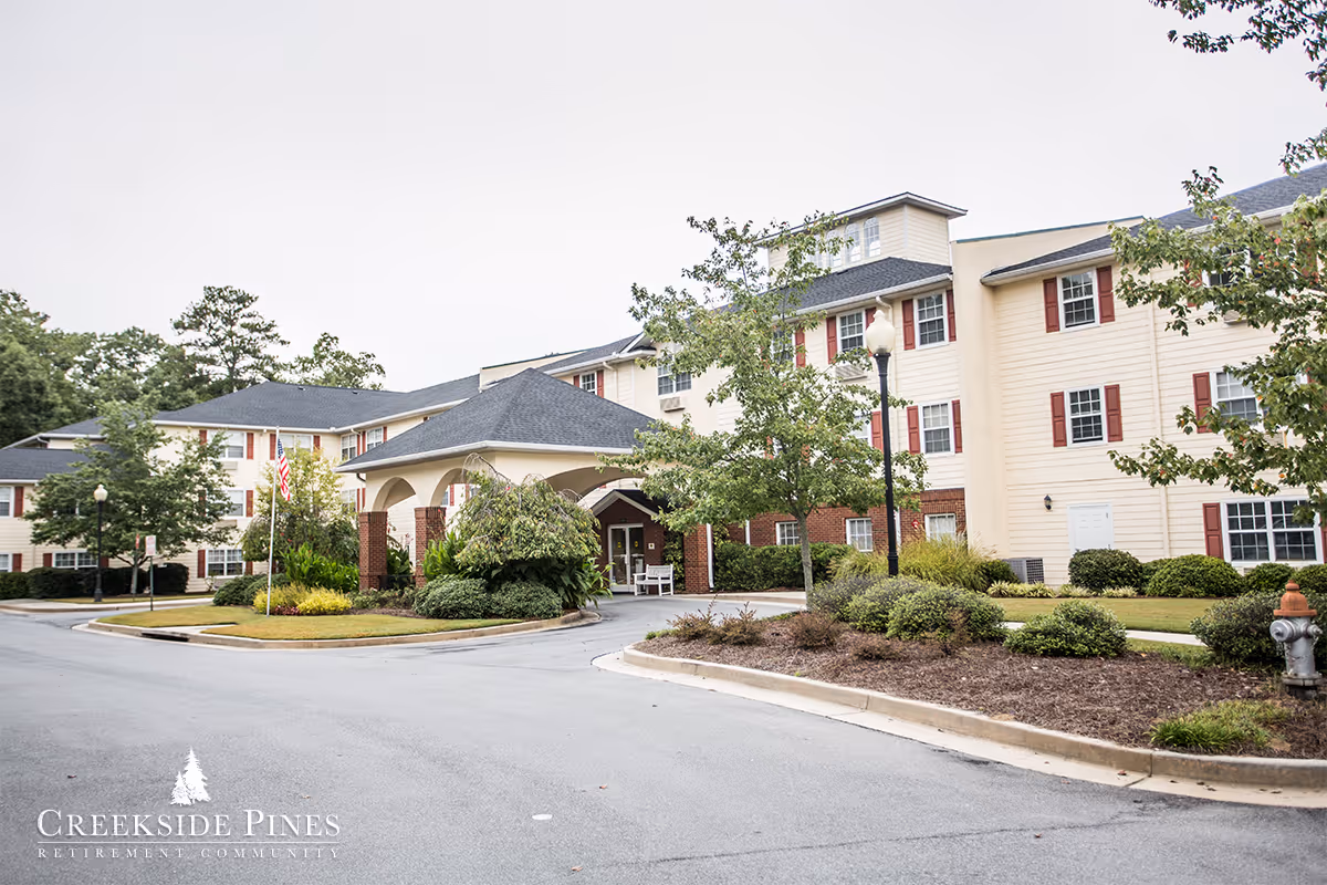 Exterior front entrance of a multi-story retirement community building with a covered porte-cochere, driveway and landscaped grounds.