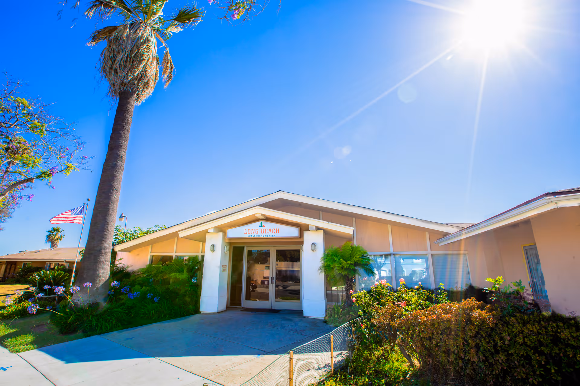 Exterior view of Long Beach Healthcare Center building entrance with a clear blue sky, palm trees, and an American flag on the left side.